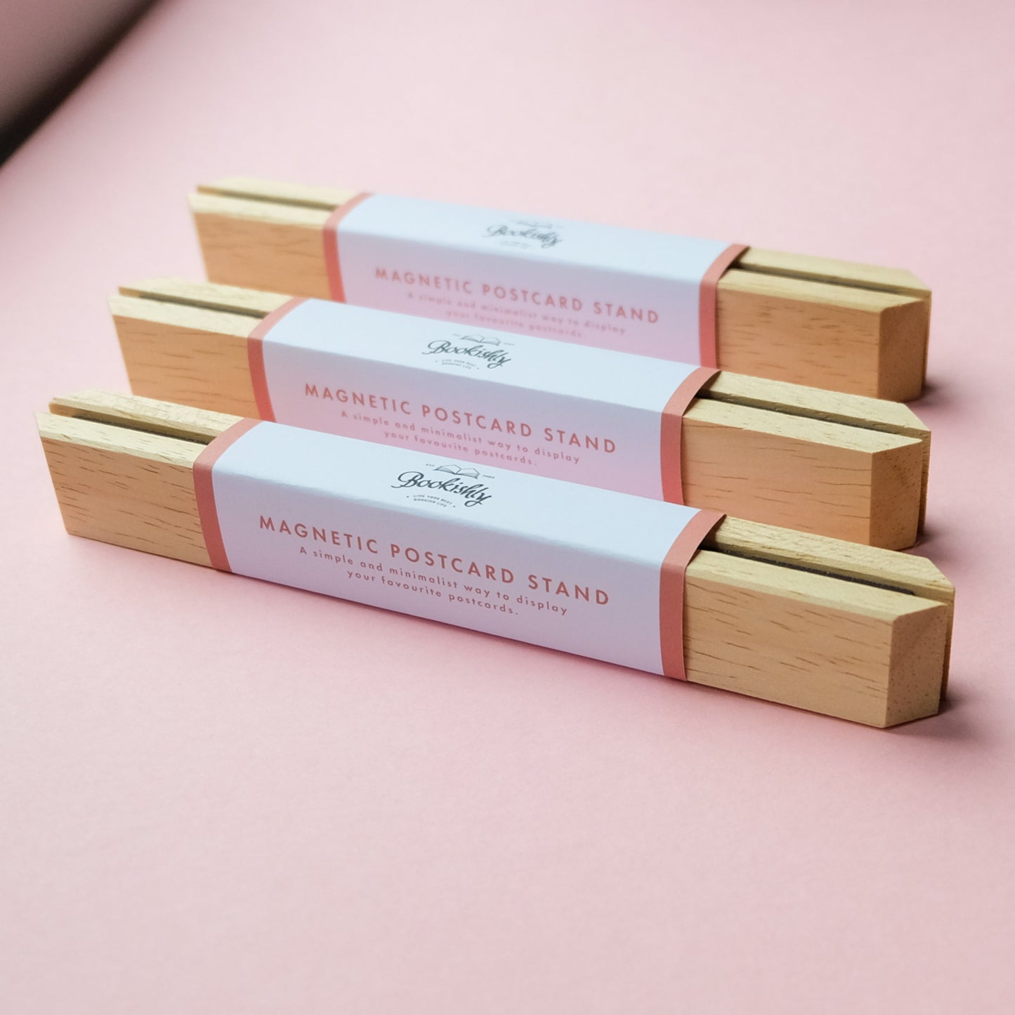 Three wooden magnetic postcard stands with packaging on a pink background