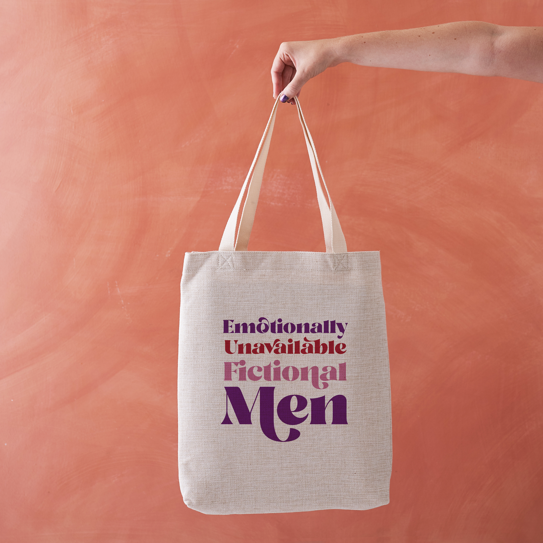 Linen-effect tote bag with the phrase “Emotionally Unavailable Fictional Men” in red and purple text, held against a warm terracotta background.