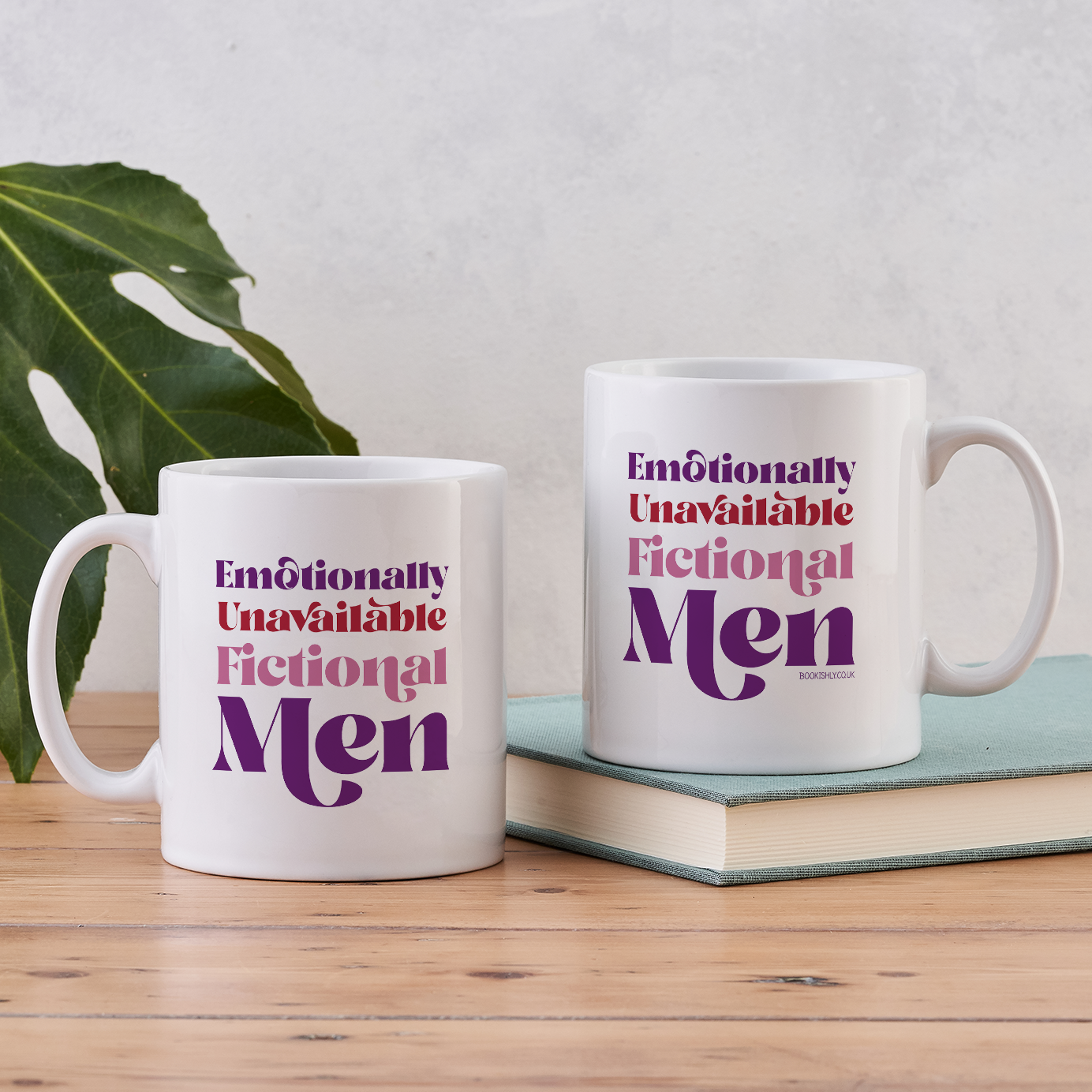 White ceramic mug with “Emotionally Unavailable Fictional Men” printed in red and purple lettering, styled beside a book on a wooden table.