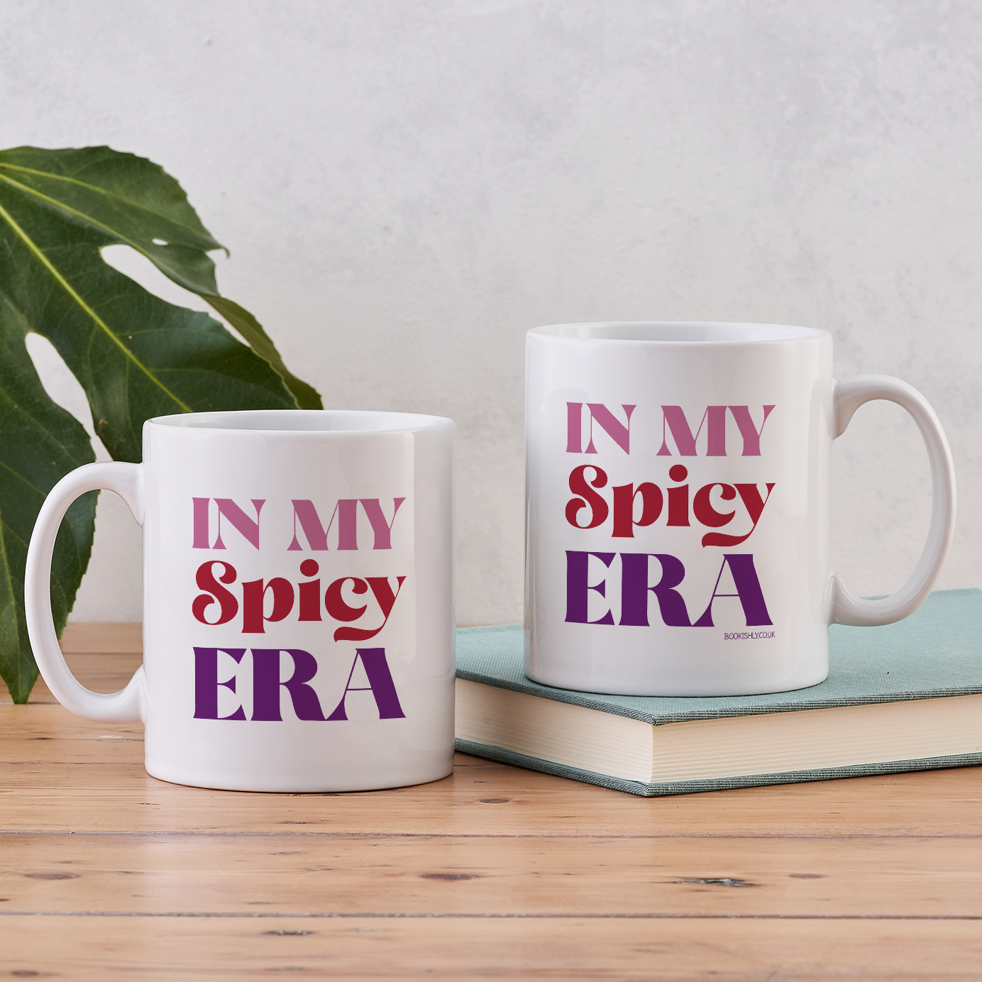 White ceramic mug with “In My Spicy Era” printed in red and purple lettering, styled on a wooden table with a green leaf.