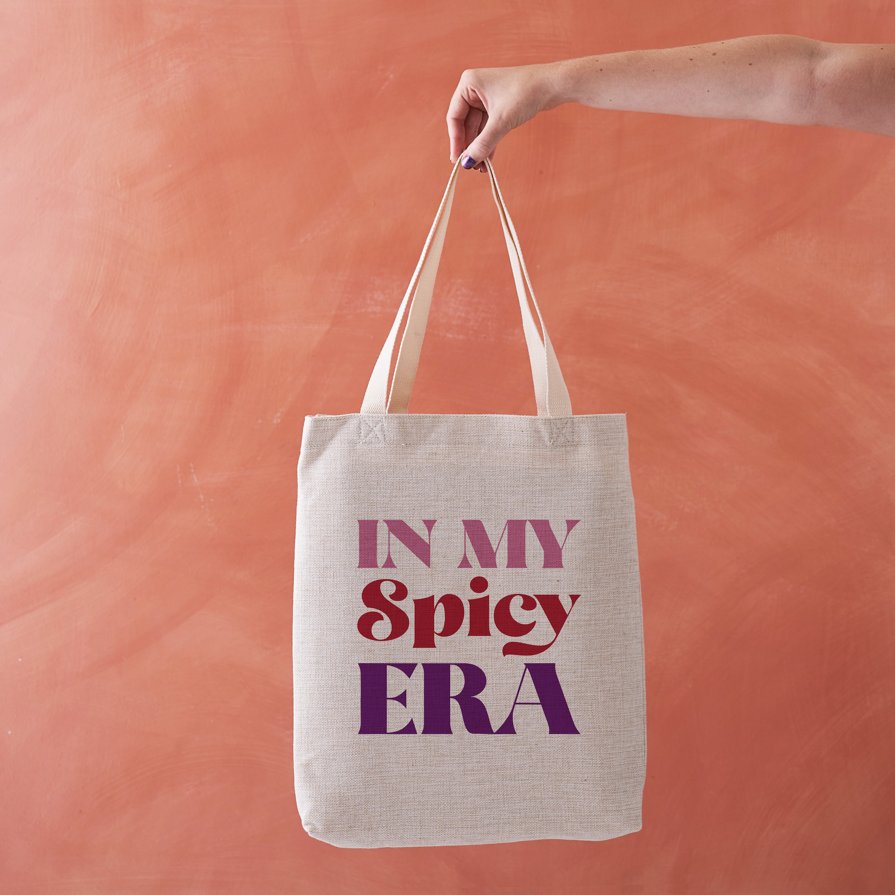 Linen-effect tote bag with the phrase “In My Spicy Era” in red and purple lettering, held against a terracotta background.