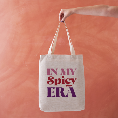 Linen-effect tote bag with the phrase “In My Spicy Era” in red and purple lettering, held against a terracotta background.
