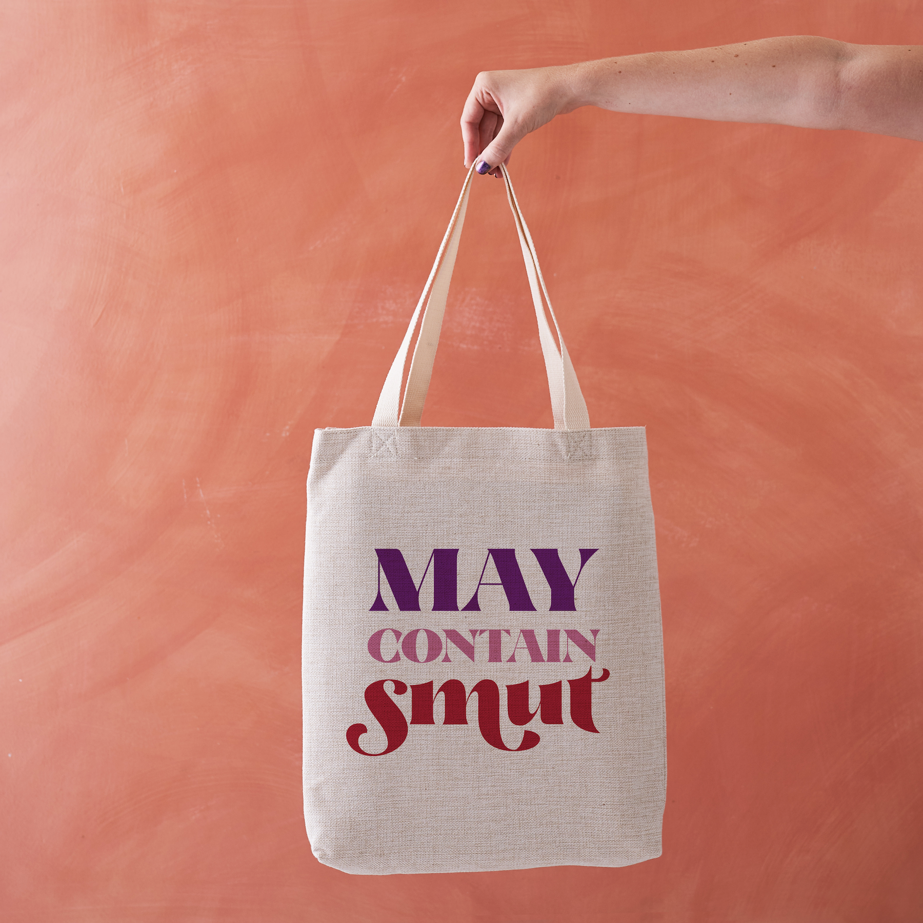 Linen-effect tote bag with the phrase “May Contain Smut” in red and purple lettering, held against a warm terracotta background.