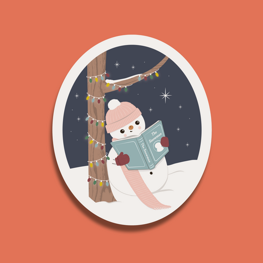 Oval sticker showing a snowman leaning against a tree with fairy lights and reading the book The Snowman