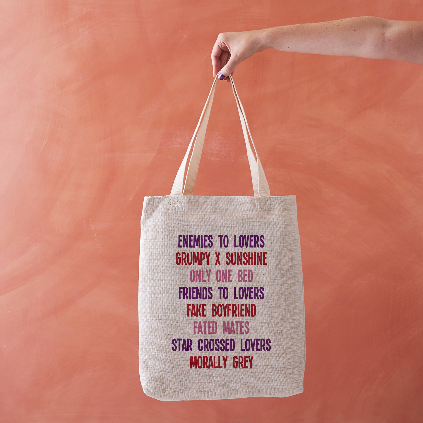 Linen-effect tote bag printed with a list of romance tropes in red, pink, and purple text, held against a warm terracotta background.