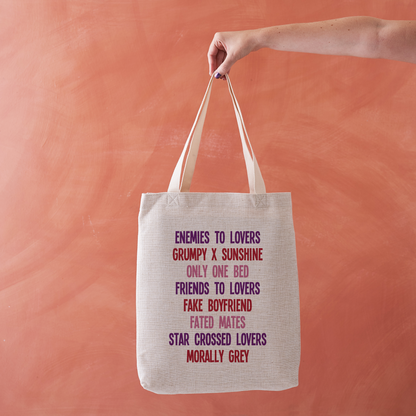 Linen-effect tote bag printed with a list of romance tropes in red, pink, and purple text, held against a warm terracotta background.