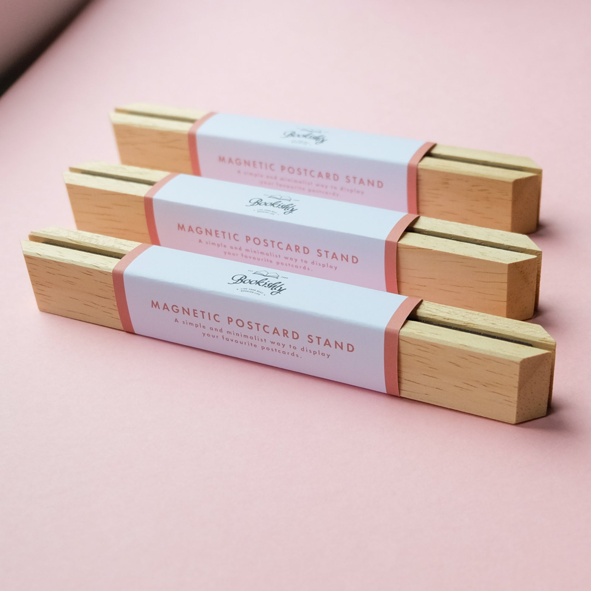 Three wooden magnetic postcard stands with packaging on a pink background
